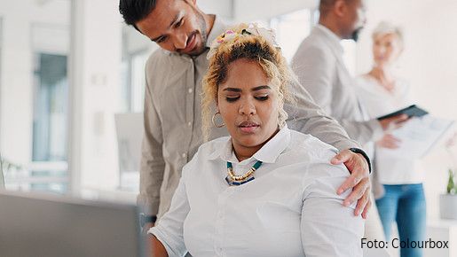 Foto zum Thema sexuelle Belästigung am Arbeitsplatz: Zu sehen ist eine Frau, die am Schreibtisch im Büro sitzt, und von einem Mann an der Schulter berührt wird. Ihr Blick verrät, dass es ihr nicht gefällt.