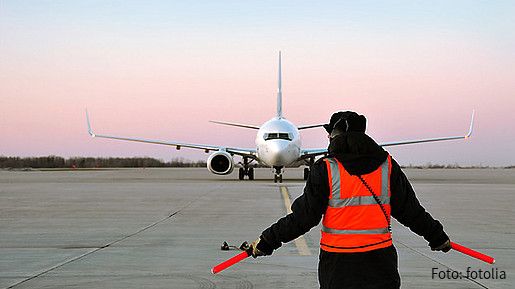 Ein Fluglotse in einer roten Warnweste steht mit dem Rücken zur Kamera. Weit vor ihm ist ein Flugzeug zu sehen, das auf der Flugbahn steht.